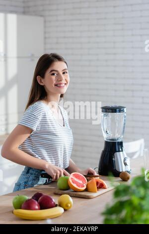 femme joyeuse regardant l'appareil photo tout en coupant des fruits frais premier plan flou Banque D'Images