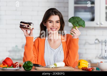 une femme gaie qui choisit entre gâteau et brocoli frais tout en souriant à l'appareil photo Banque D'Images