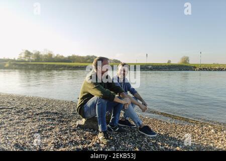 Le père et le fils passent du temps libre assis au bord de la rivière contre ciel dégagé Banque D'Images
