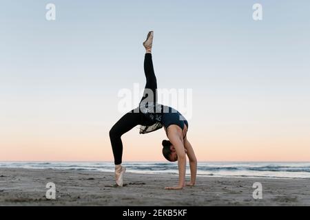 Danseuse de ballet flexible se pliant vers l'arrière tout en s'exerçant à la plage contre le ciel Banque D'Images Danseuse de ballet flexible se pliant vers l'arrière tout en s'exerçant à la plage contre le ciel Banque D'Images