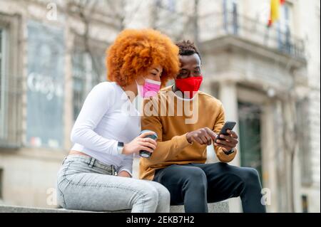 Couple portant un masque facial avec un téléphone portable lorsqu'il est assis ville Banque D'Images