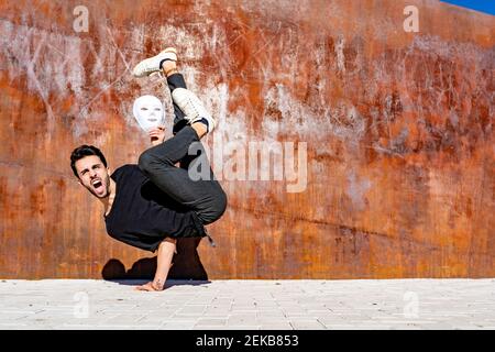 Jeune homme avec masque blanc criant tout en faisant la main contre mur brun Banque D'Images