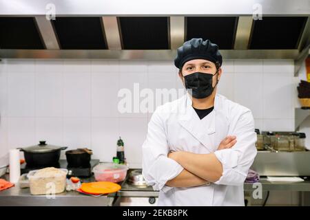 Jeune chef masculin en masque de protection avec les bras croisés dans la cuisine commerciale Banque D'Images