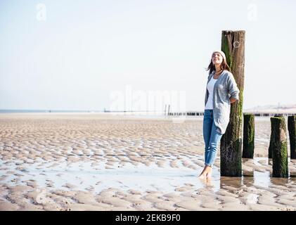Jeune femme avec les yeux fermés, penchée sur un poteau en bois à plage Banque D'Images