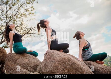 Des femmes flexibles assises avec des jambes croisées tout en faisant de l'exercice d'étirement sur rochers Banque D'Images