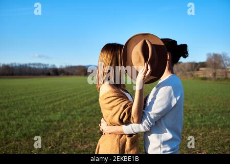 Couple hétérosexuel embrassant tandis que la petite amie couvrant les visages avec le chapeau contre ciel bleu clair Banque D'Images