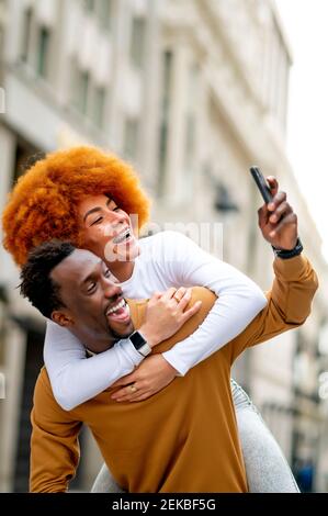 Homme prenant le selfie par téléphone mobile tout en faisant un porcgysoutenant la femme ville Banque D'Images