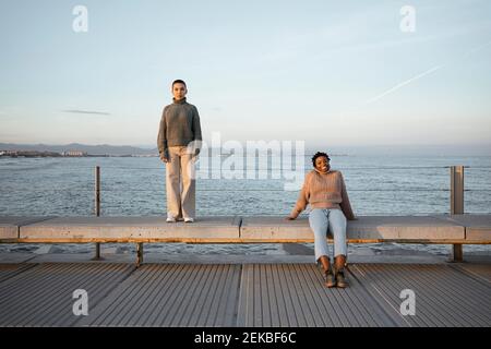 Triste femme debout à distance avec un ami heureux assis sur banc contre la mer Banque D'Images