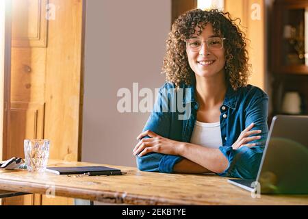 Bonne jeune femme d'affaires avec les bras croisés par ordinateur portable à la maison bureau Banque D'Images