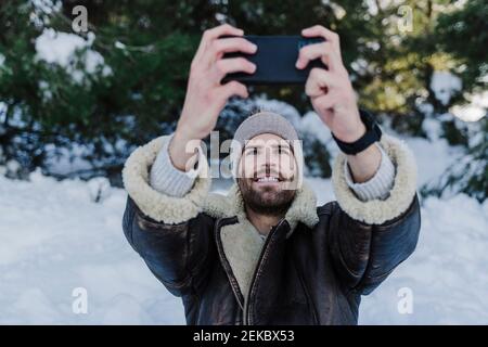 Homme souriant dans des vêtements chauds emportant le selfie à travers un smartphone en hiver Banque D'Images