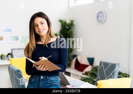 Femme souriante, étudiant tenant un ordinateur portable et un stylo tout en restant debout séjour Banque D'Images