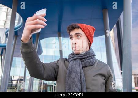 Jeune homme en tricot tout en emportant le selfie à travers un smartphone en ville Banque D'Images