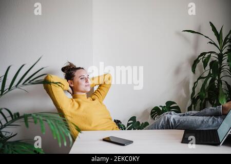 Femme souriante avec les mains derrière la tête se détendant tout en étant assise table à la maison Banque D'Images