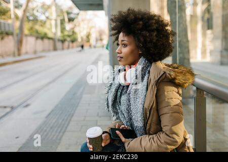 Femme afro avec téléphone intelligent et tasse à café jetable contemplant à la gare routière en hiver Banque D'Images