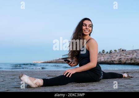 Danseuse de ballet souriante regardant l'épaule tout en faisant la fente de jambe à la plage contre un ciel dégagé Banque D'Images