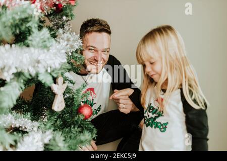 Fille montrant l'ornement de Noël à l'appareil-photo tandis que la famille décorant l'arbre de Noël dans la maison Banque D'Images