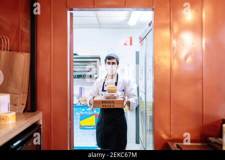 Un chef masculin transportant des boîtes à pizza et un récipient à nourriture pendant son travail au restaurant pendant une pandémie Banque D'Images