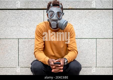 Jeune homme portant un masque de gaz de pollution assis avec un téléphone portable contre le mur Banque D'Images