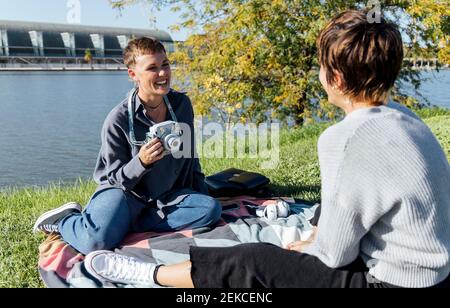 Femme souriante avec un appareil photo regardant un ami assis contre rivière au parc Banque D'Images