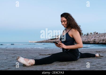 Danseuse flexible utilisant un téléphone portable tout en faisant des fractionnements à plage contre ciel clair Banque D'Images