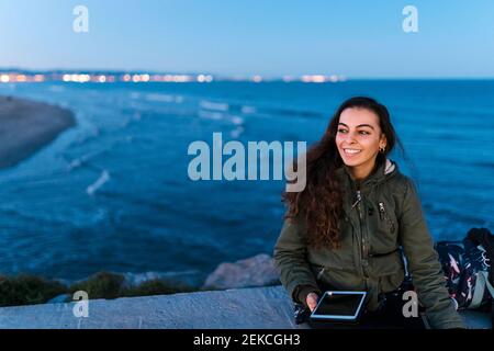 Femme souriante avec une tablette numérique qui regarde loin tout en étant assise mur de retenue contre la mer Banque D'Images