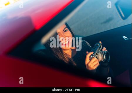 Jeune femme souriante avec appareil photo vintage dans la voiture vue à travers pare-brise par temps ensoleillé Banque D'Images