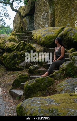 Belle jeune femme caucasienne assise avec une robe verte un escalier à l'extérieur plein de rochers et d'arbres avec mousse