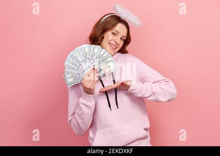 Portrait d'une adolescente à cheveux bouclés à succès avec halo sur la tête montrant un tas de billets de banque en dollars et sincèrement souriant, campagne de charité Banque D'Images