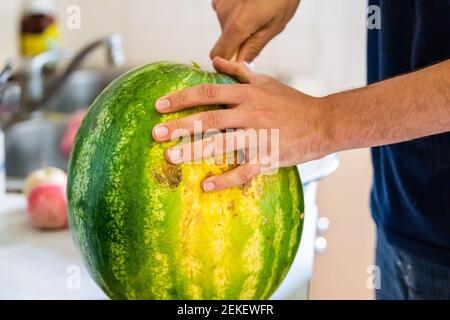 Gros plan du jeune homme à la maison tenant couper en deux la pastèque avec couteau et fruit a un champ jaune indiquant douceur Banque D'Images