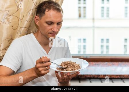 Jeune homme assis sur une chaise par des stores de seuil de fenêtre vintage En Ukraine ou en Russie, manger du sarrasin à partir d'une assiette à portée de main avec la fourche Banque D'Images