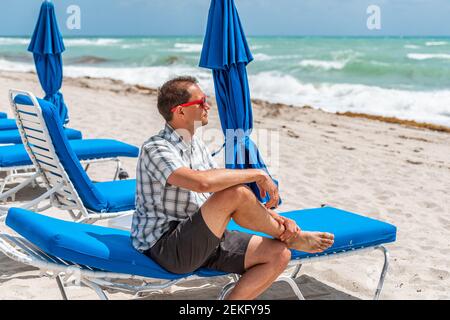 Jeune homme hipster millénaire en lunettes de soleil rouges sur la plage le jour ensoleillé à Miami, Floride assis sur des chaises de plage bleues, parasols près de l'océan Atlantique g Banque D'Images