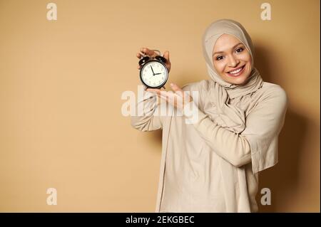 Femme arabe heureuse avec tête couverte dans hijab tenant l'alarme horloge dans les mains et sourire mignon avec un sourire en dents de souris à caméra en se tenant contre un beige b Banque D'Images