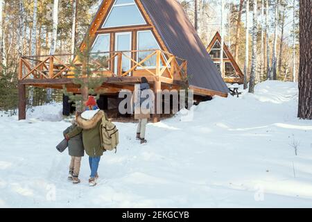 Jeune homme avec grand sac à dos se déplaçant vers la maison de campagne pendant sa femme et leur petite fille en hiver chaud après lui Banque D'Images