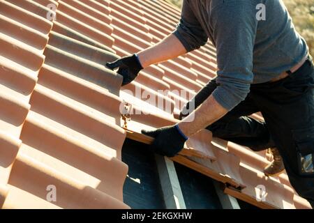 Gros plan des mains du travailleur installant des tuiles de toiture en céramique jaune montées sur des panneaux en bois recouvrant le toit d'un bâtiment résidentiel en construction. Banque D'Images