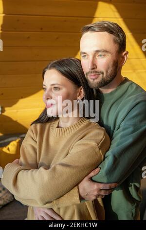 Jeune mari affectueux qui embrasse sa femme en se tenant devant grande fenêtre et profiter du week-end dans leur maison de campagne Banque D'Images