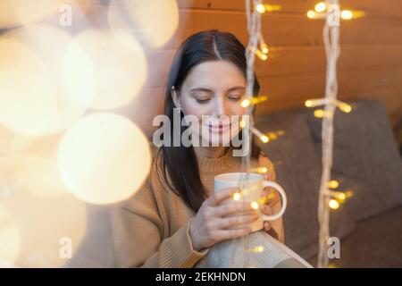 Jeune femme sereine brune avec une tasse de thé chaud assis sur le canapé par une grande fenêtre de la maison de campagne en hiver week-end et repos Banque D'Images