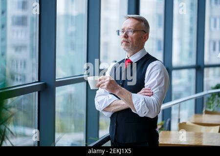 Sourire heureux directeur général pense à son développement de carrière réussi tout en se tenant avec une tasse de café dans sa main dans son bureau près de la Banque D'Images