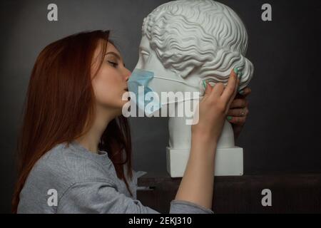 Jeune fille aux cheveux rouges sur fond noir dans un t-shirt gris, embrassant avec un buste en plâtre dans un masque médical protecteur Banque D'Images