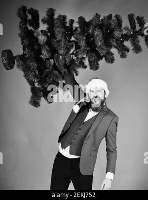 Manager avec barbe se prépare pour Noël. Concept d'affaires et de vacances d'hiver. Homme en costume élégant et chapeau de père Noël sur fond rouge. Un homme d'affaires au visage déchaînant tient un arbre de Noël chauve. Banque D'Images