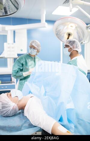 Jeune homme patient dans un masque de protection et des vêtements de tête couchés table d'opération pendant que deux chirurgiens se préparent à l'opération Banque D'Images