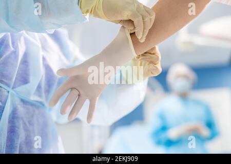 Assistant dans une combinaison mettant un gant de protection à portée de main chirurgien tout en se préparant à l'opération sur le fond de la femme dans vêtements de travail Banque D'Images