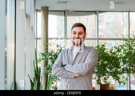 Homme caucasien souriant debout près de la fenêtre dans le bureau. Jeune homme intelligent barbu avec succès et bras croisés. Beau homme d'affaires dans une chemise décontractée. Gratuit Banque D'Images