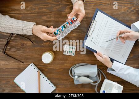 Vue d'ensemble des mains de l'homme âgé malade tenant un contenant avec pilules sur la table pendant la consultation médicale avec le médecin remplissant document Banque D'Images