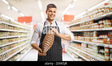 Jeune homme assistant de magasin portant un tablier et tenant un pain de pain brun dans un supermarché Banque D'Images