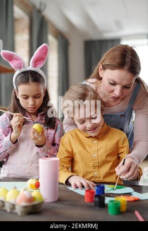 Mère aidant son petit fils à peindre une image pendant Sa fille peignant des œufs qu'ils préparent pour Pâques Banque D'Images