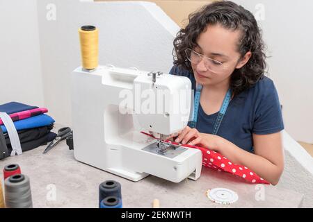 portrait d'une femme latine attrayante utilisant une machine à coudre, en plus des ciseaux, des fils, du tissu, faisant des vêtements à la maison comme petite femme d'affaires Banque D'Images