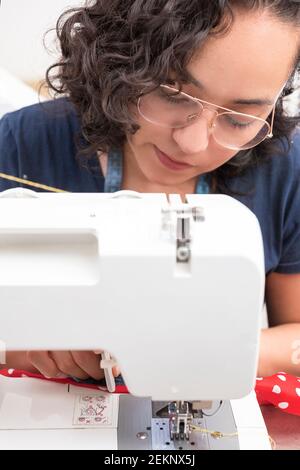 portrait d'une femme latine attrayante utilisant une machine à coudre, en plus des ciseaux, des fils, du tissu, faisant des vêtements à la maison comme petite femme d'affaires Banque D'Images