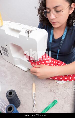 portrait d'une femme latine attrayante utilisant une machine à coudre, en plus des ciseaux, des fils, du tissu, faisant des vêtements à la maison comme petite femme d'affaires Banque D'Images