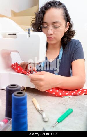 portrait d'une femme latine attrayante utilisant une machine à coudre, en plus des ciseaux, des fils, du tissu, faisant des vêtements à la maison comme petite femme d'affaires Banque D'Images