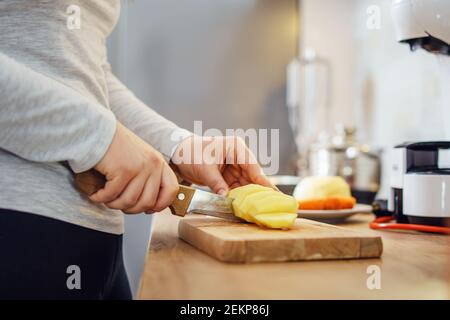 Vue latérale sur les mains d'une femme inconnue avec couteau montez dans la cuisine en coupant des morceaux de pomme de terre pour le soupe végétalienne ou végétarienne cuisant un repas sain Banque D'Images